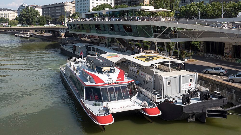 Modern passagerarfärja förtöjd bredvid ett flytande kontor på en flod i en urban stadsbild med en bro i bakgrunden