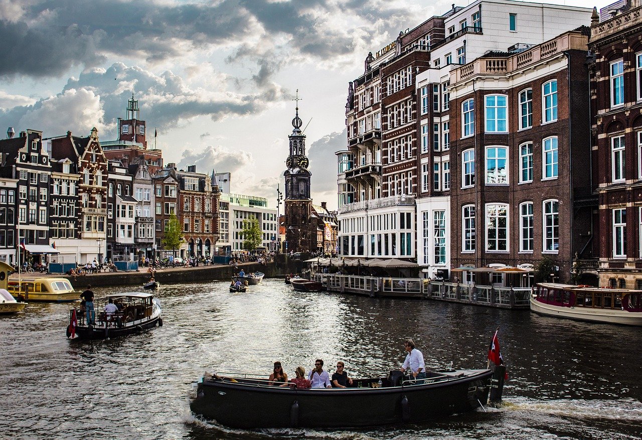 Turister som njuter av båtturer på en kanal omgiven av historiska byggnader i Amsterdam under en molnig himmel