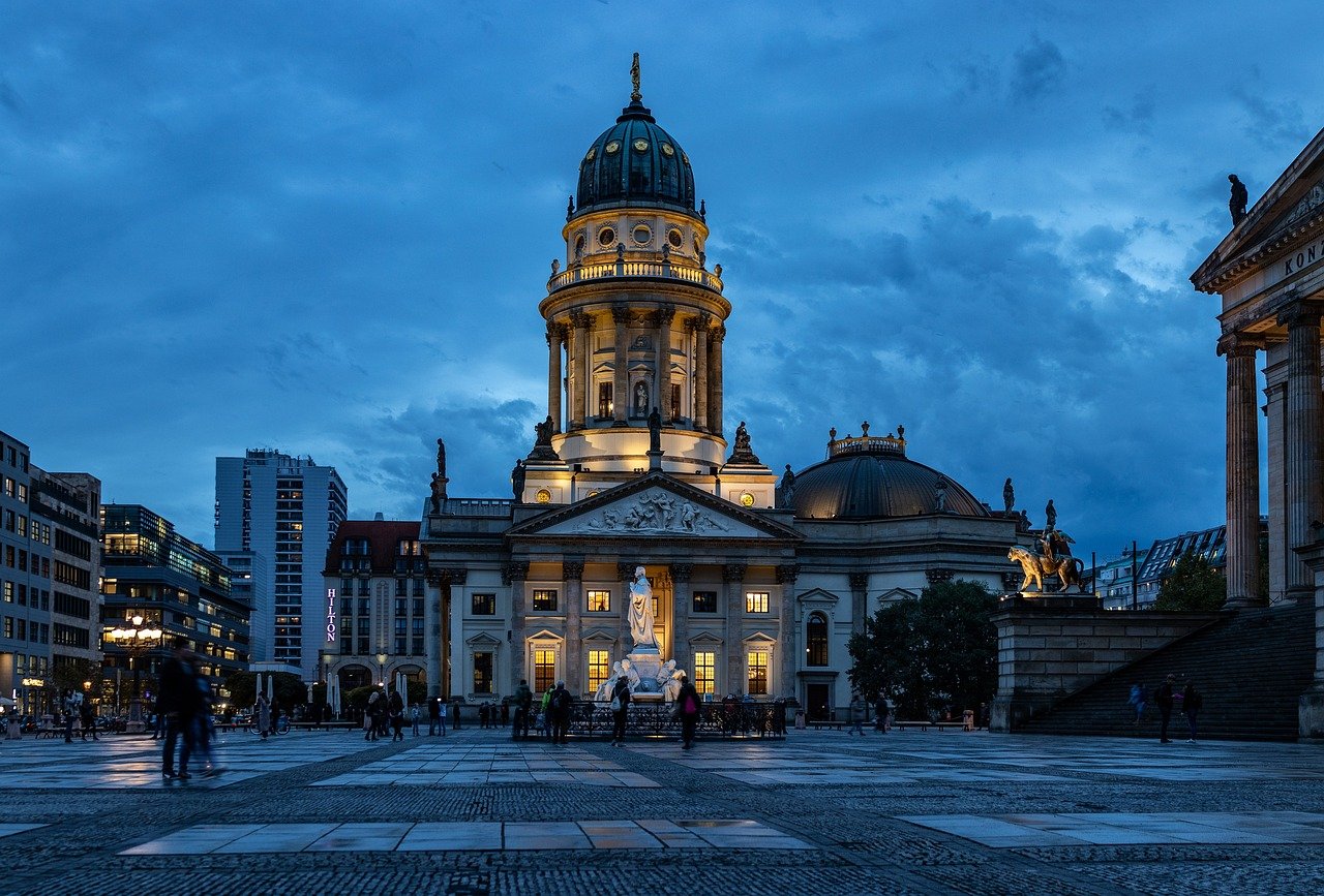 Upplyst historisk katedral med kupol och statyer på stadsplats i skymningen med människor som promenerar runt