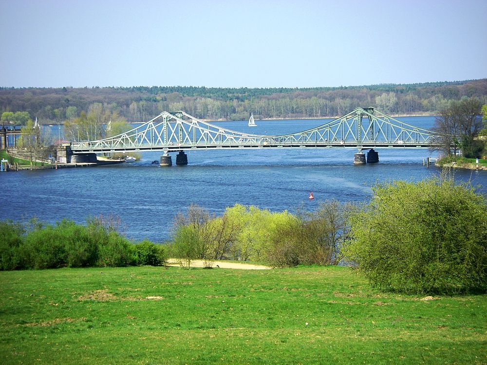 Ståltrussbro över flod med grönska Scenisk vy av en ståltrussbro över en bred flod med grönt gräs och träd i förgrunden under klarblå himmel