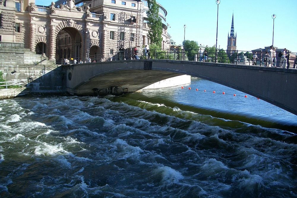 Stenvalvsbro över flod med historiska byggnader Stenvalvsbro över turbulent flodvatten med historiska byggnader och kyrktorn i bakgrunden under klar himmel