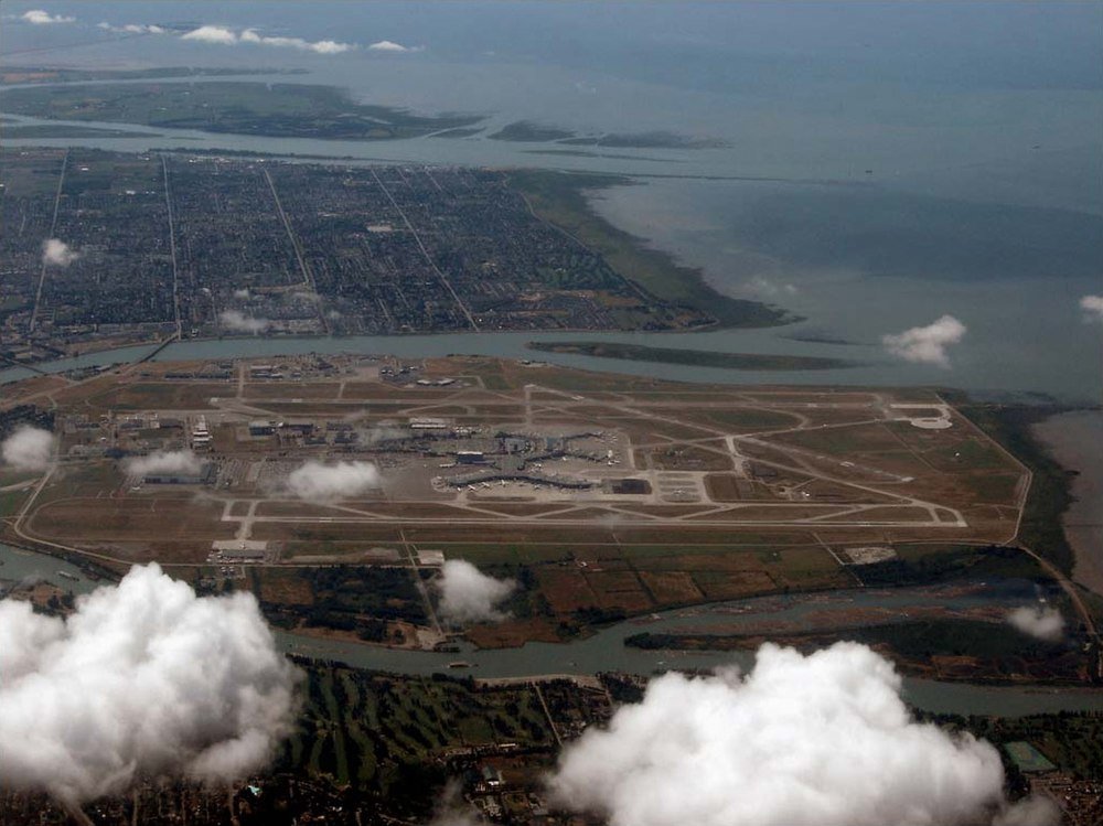 Flygfoto av öflygplats med landningsbanor och terminaler Flygfoto av en stor flygplats på en ö omgiven av vatten och urbana områden med landningsbanor och terminaler synliga