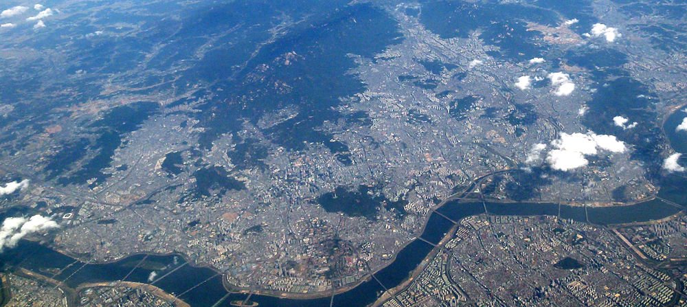 Flygfoto av en stor urban stad med floder, broar och spridda moln över bergig terräng