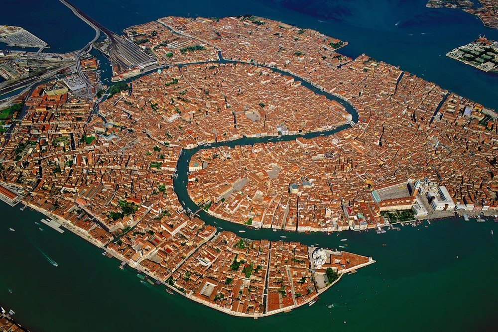 Flygfoto av Venedig Italien som visar historiska byggnader och kanaler omgivna av vatten, vilket framhäver stadens unika arkitektur.