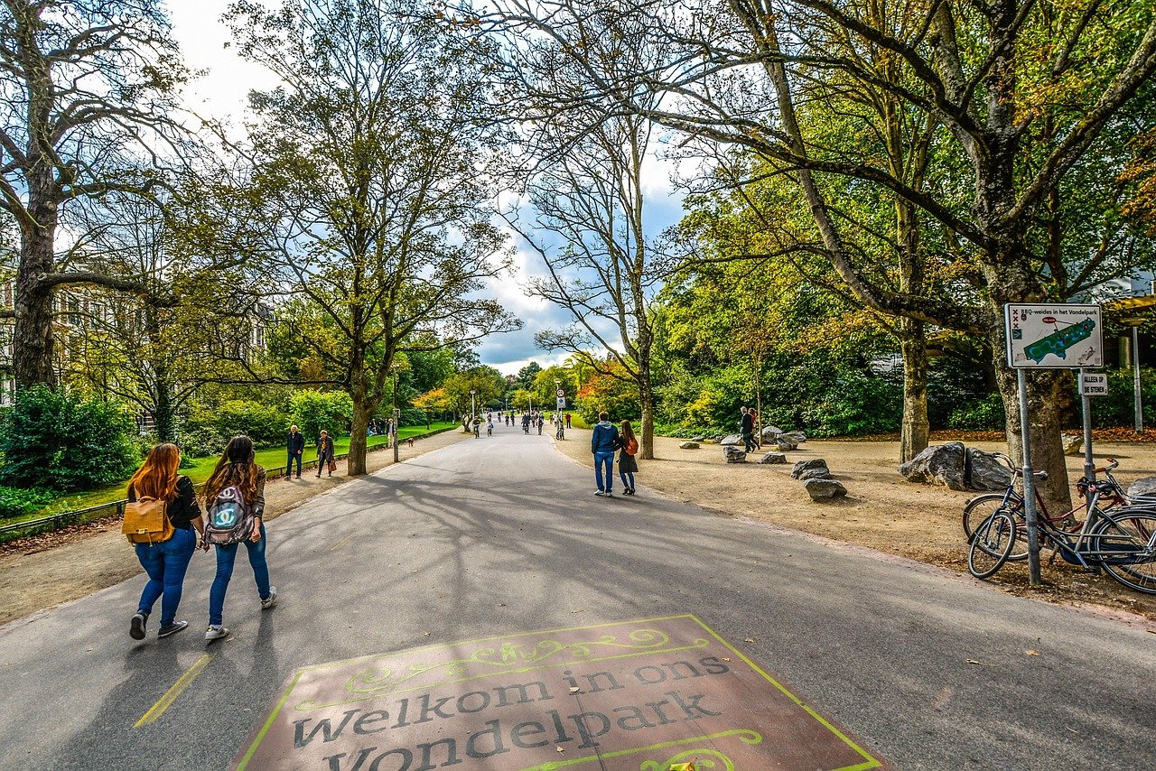 Människor som går och cyklar i Vondelpark Amsterdam med träd och skyltar som välkomnar besökare till parken