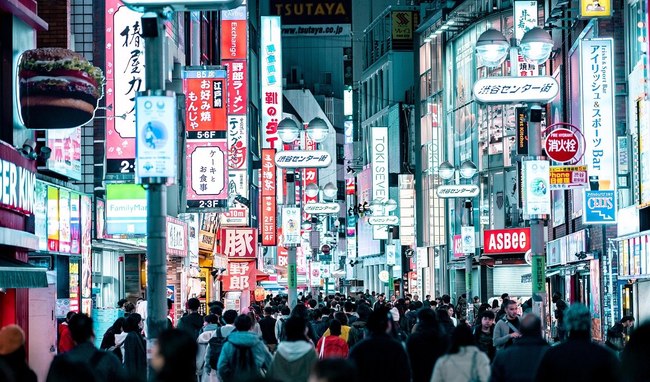 Trång urban gata i Tokyo på natten med ljusa neonskyltar och olika människor som går under upplysta annonser
