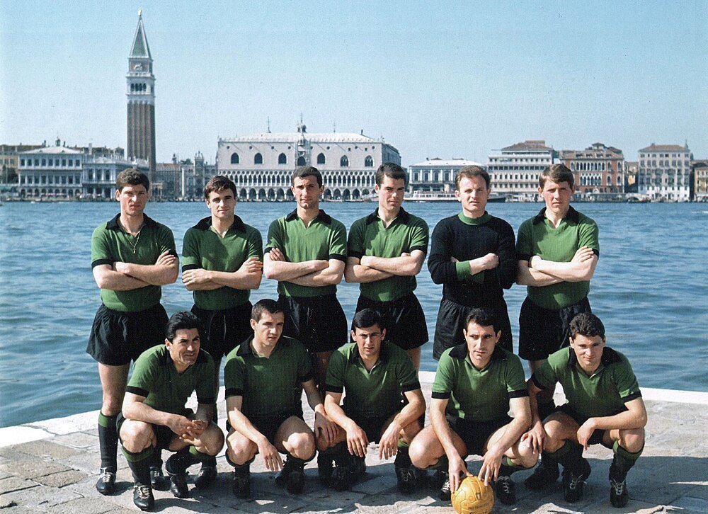 Vintage fotbollslag poserar vid venetianskt vattnet Vintage fotbollslag i gröna uniformer poserar vid vattnet med historiska venetianska byggnader och klocktorn i bakgrunden