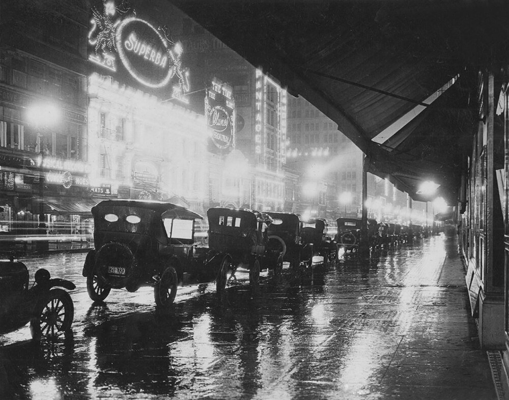 Vintage svartvitt foto av tidiga 1900-talets bilar parkerade på en regnig stadsgata med ljusa neonteaterskyltar