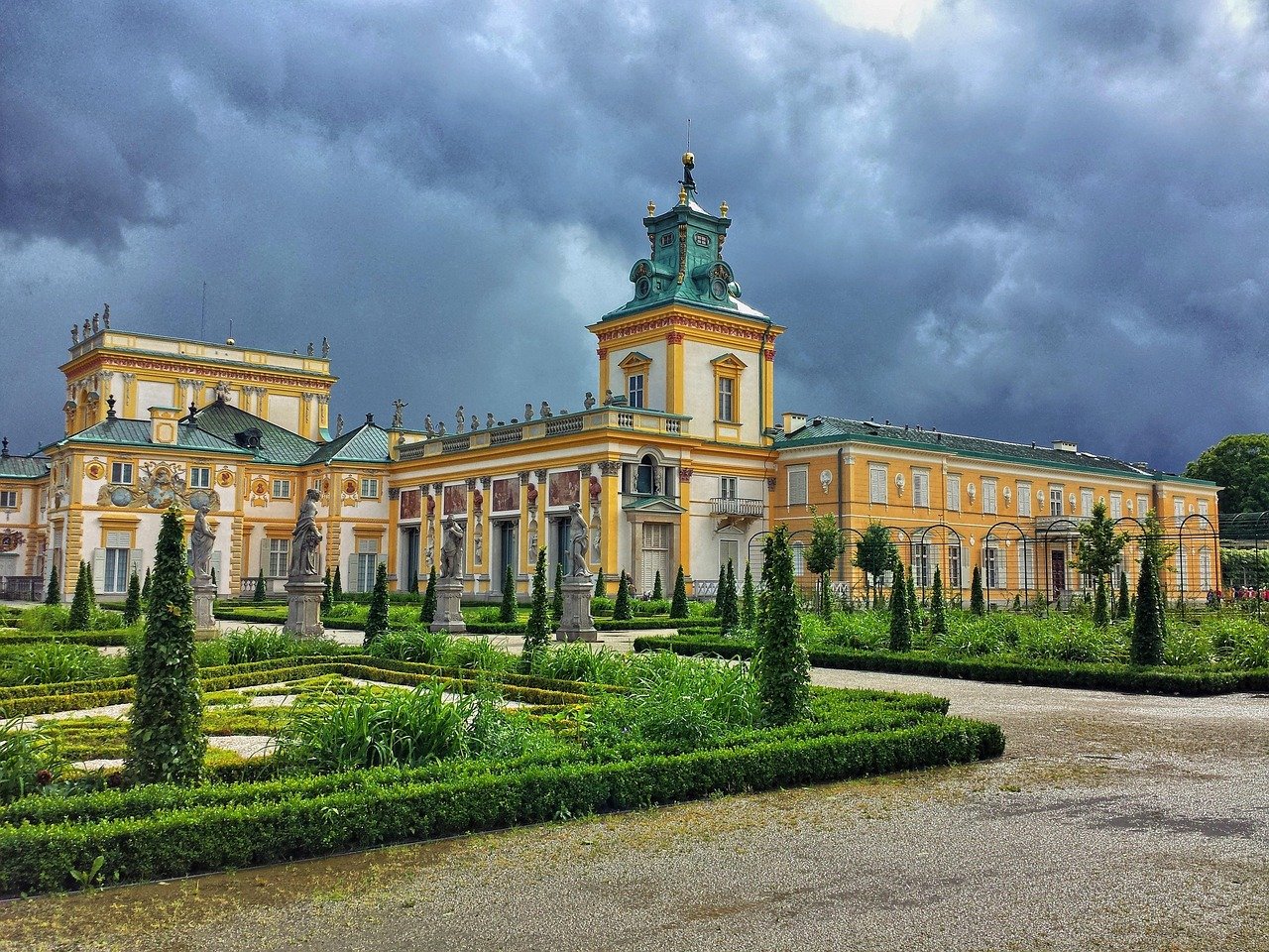 Barockstil palats med utsmyckade statyer och välskötta trädgårdar under en dramatisk molnig himmel