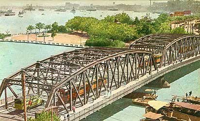 Historisk ståltrussjärnvägsbro över flod med tåg Historisk ståltrussjärnvägsbro över flod med tåg och båtar, omgiven av grönska och stadsbild i bakgrunden