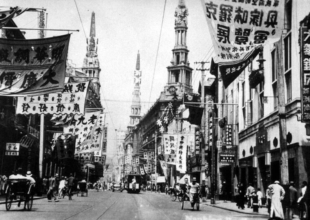 Historisk svartvit gatubild i tidigt 1900-talets Shanghai med traditionella kinesiska banderoller och rickshaws