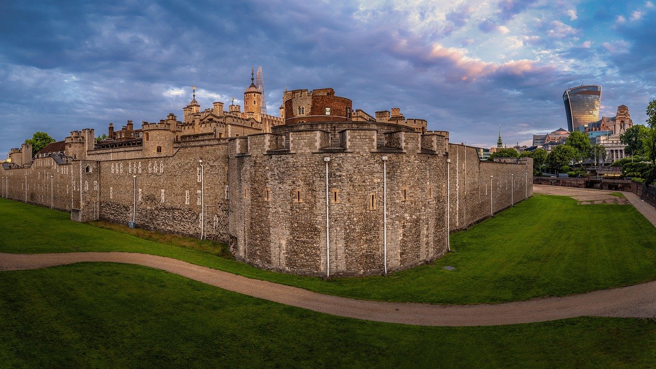 Historiska Tower of London fästning med stenväggar och modern stadssilhuett under en molnig himmel vid solnedgången