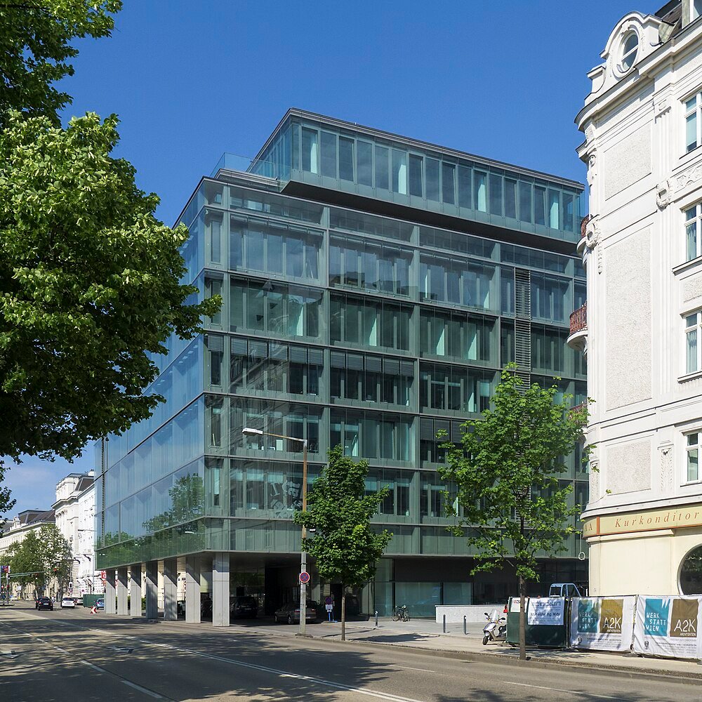 Modern kontorsbyggnad i glas med flera våningar och träd längs gatan under en klarblå himmel