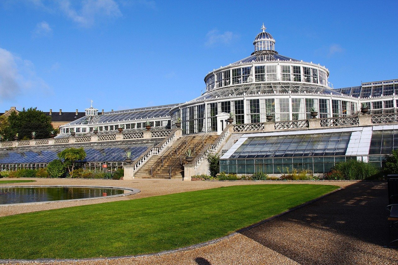 Stort historiskt växthus av glas med terrasserade glastak och stentrappor i en botanisk trädgård under blå himmel