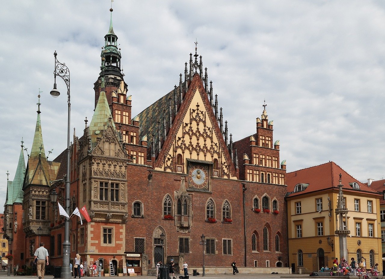 Historiskt gotiskt stadshus med utsmyckat klocktorn och röd tegelstensfasad på ett europeiskt stadstorg under molnig himmel