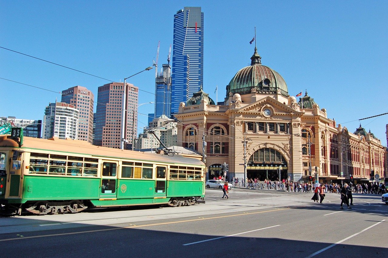 Historiska Flinders Street Station med grön spårvagn och moderna skyskrapor i Melbourne stad under klarblå himmel