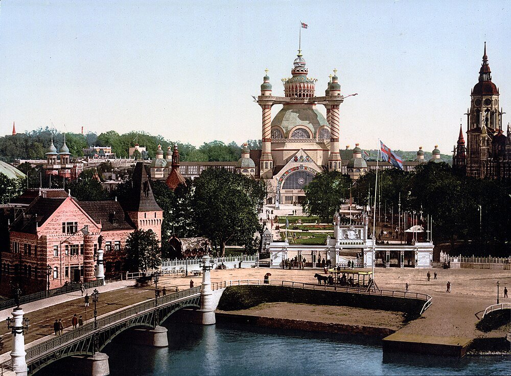 Historisk storslagen utställningshall och bro i stadspark Historisk vy av en storslagen utställningshall med utsmyckad arkitektur, en bro över vatten och omgivande byggnader i en stadspark.