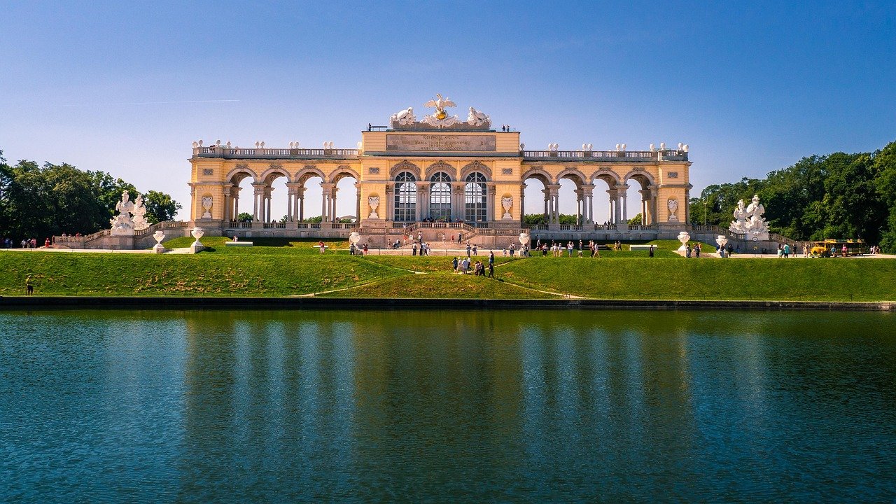 Gloriette-struktur i Schönbrunns slotts trädgårdar med blå himmel och reflekterande damm i Wien, Österrike