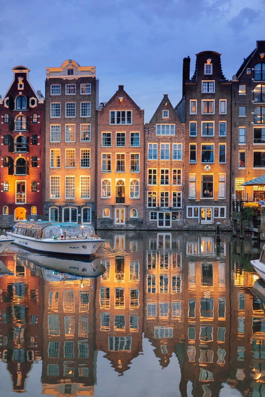 Historiska kanalhus med upplysta fönster som speglas i vattnet i Amsterdam under skymningen med en båt i förgrunden