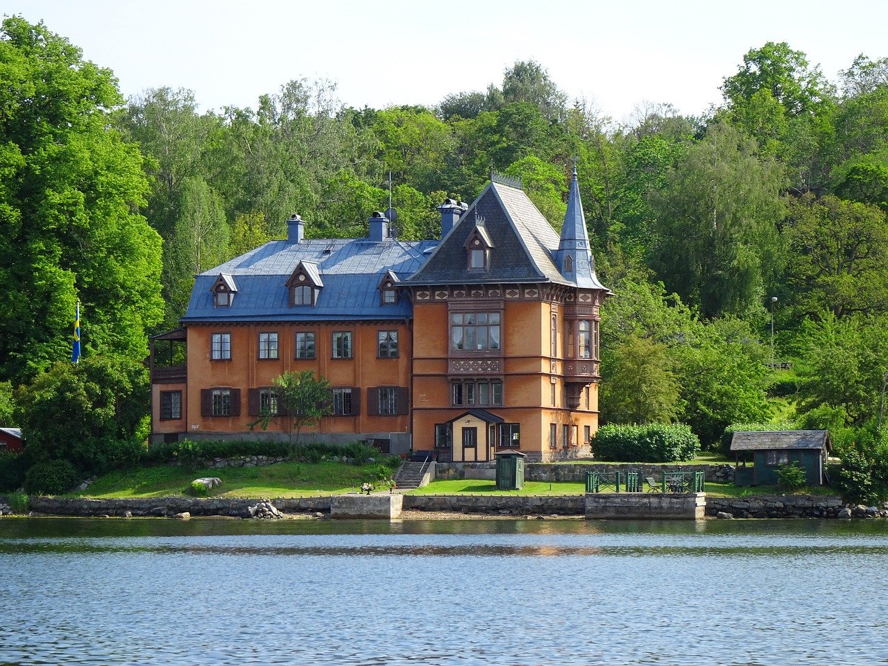 Historiskt orange hus med blått tak vid en sjö omgiven av frodiga gröna träd och klart vatten i en fridfull miljö