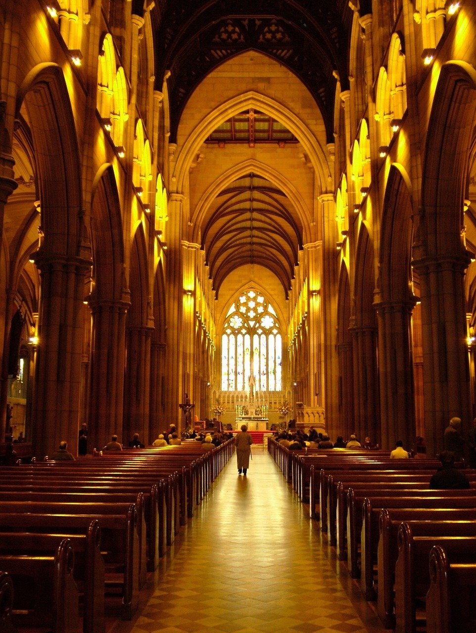 Interiörvy av en stor katedral med höga valv, träbänkar och ett lysande altare med färgade glasfönster