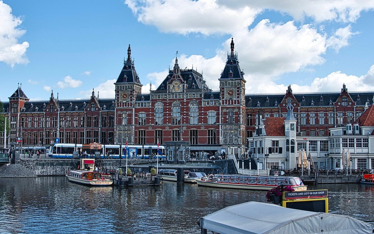 Historiska Amsterdam Centralstation med kanalbåtar och spårvagn under en klarblå himmel i Nederländerna