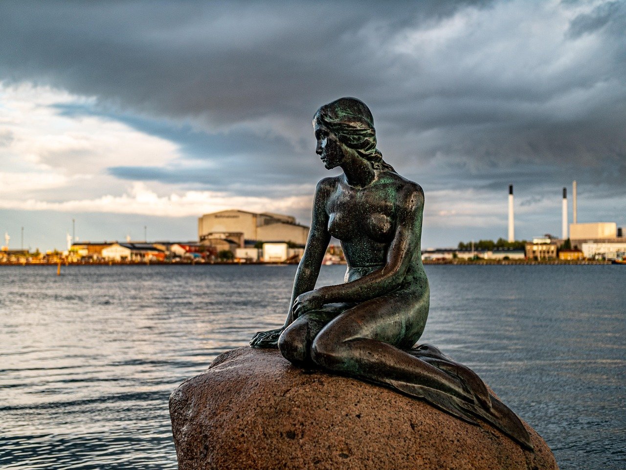 Den lilla sjöjungfrun statyn som sitter på en sten vid vattnet i Köpenhamn med industriella byggnader och molnig himmel i bakgrunden