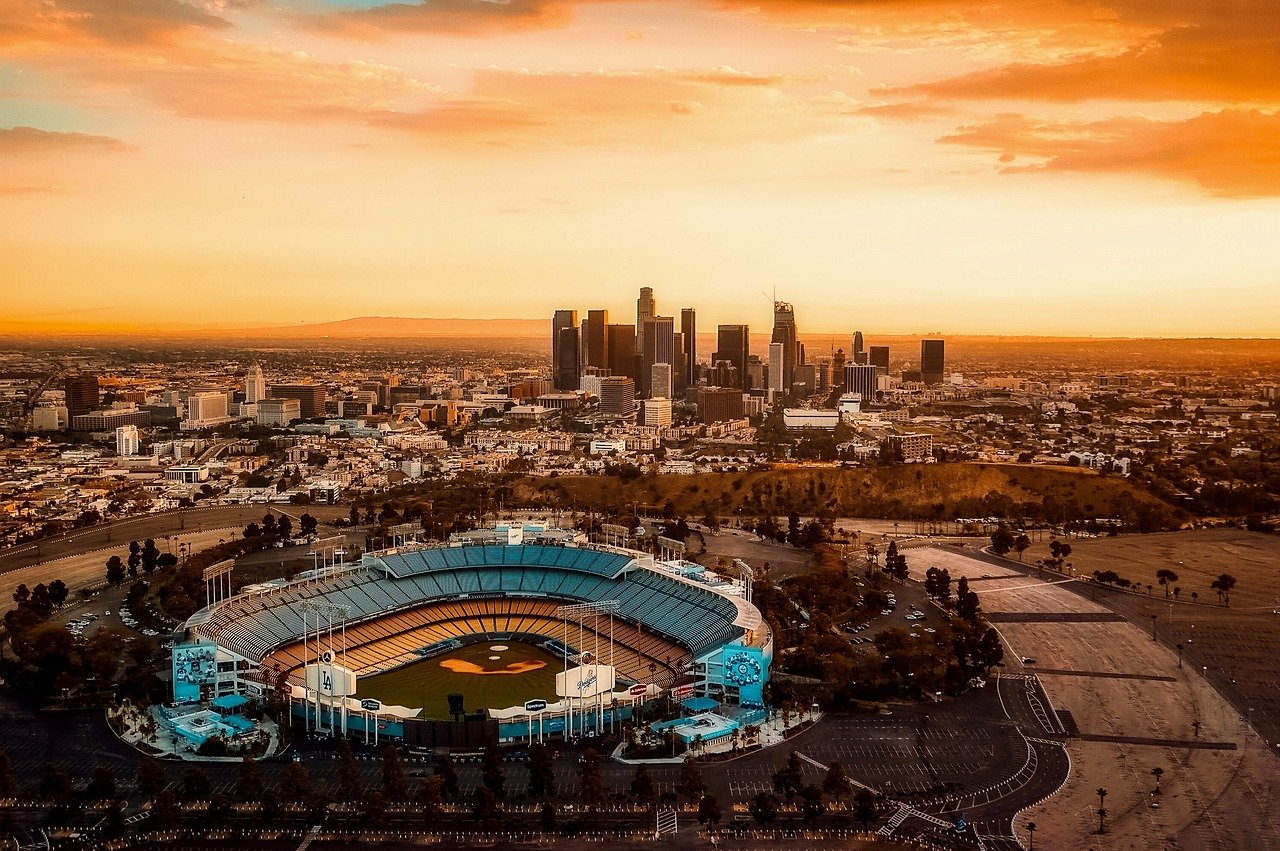 Flygfoto av Dodger Stadium med Los Angeles stadssilhuett vid solnedgången, som visar basebollplan och stadsmiljö.