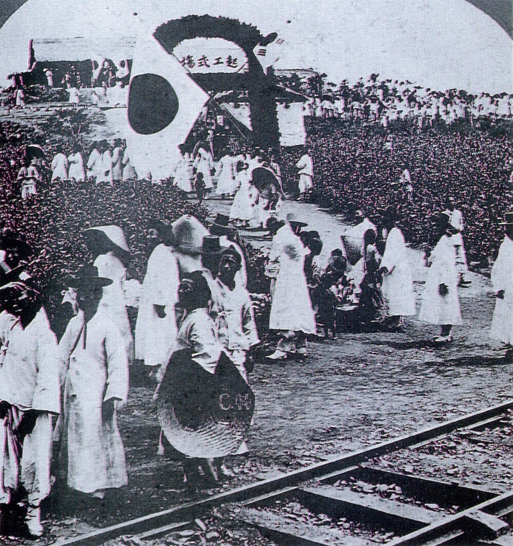 Historiska koreanska arbetare nära järnväg och väderkvarn Historiskt svartvitt foto av koreanska arbetare i traditionella kläder samlade nära järnvägsspår och en väderkvarn.