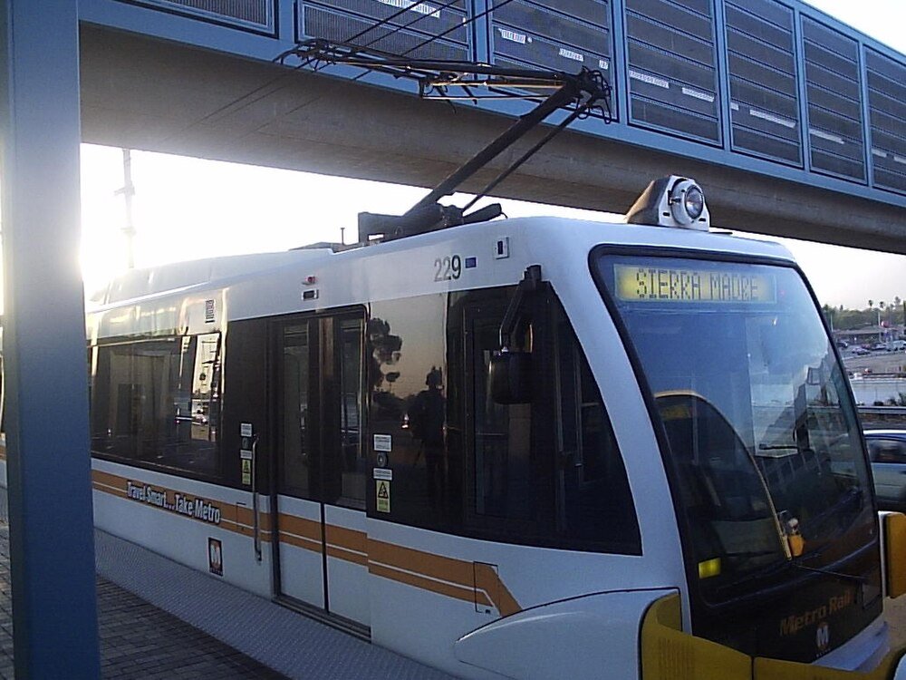 Metro Rail-tåg vid station med Sierra Madre-skylt Modern vit och orange Metro Rail-tåg vid stationsplattform med överliggande elektriska ledningar och Sierra Madre-destinationsskylt