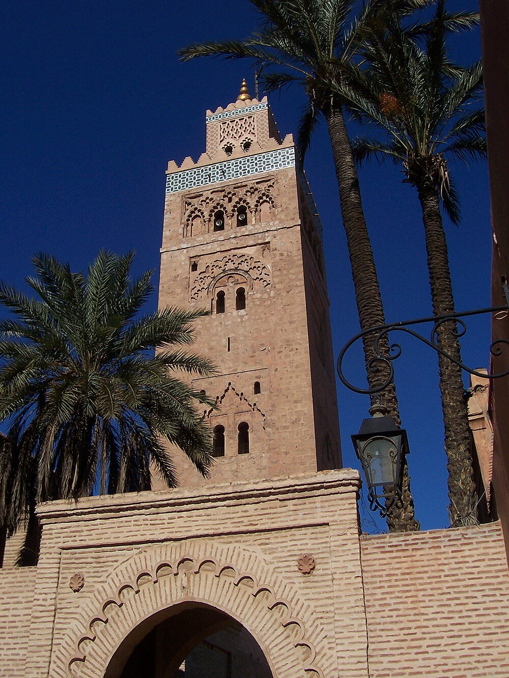 Historiskt moskétorn med palmer i Marrakech Högt historiskt moskétorn med intrikata stenristningar och palmer under klarblå himmel i Marrakech Marocko