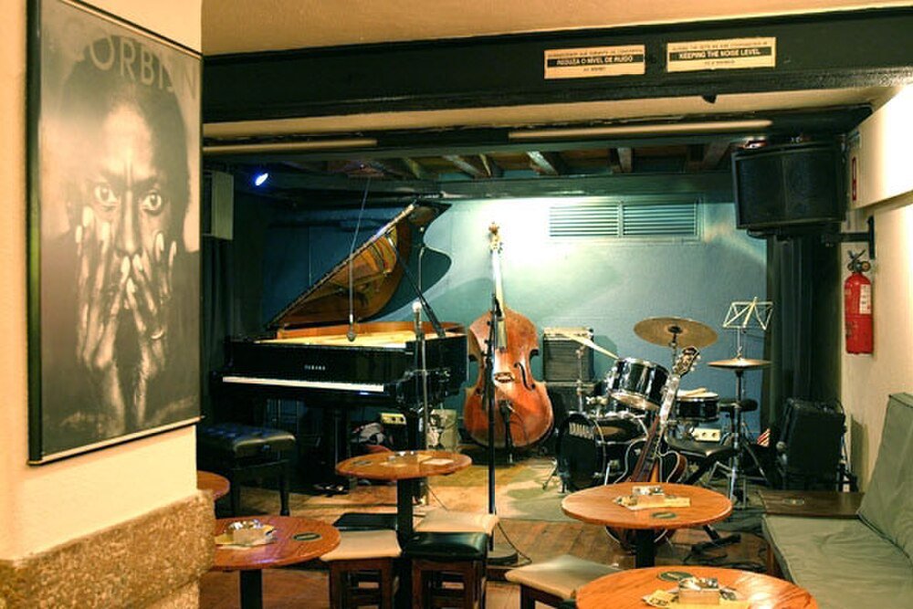 Jazzklubbscen med instrument och sittplatser Mysig jazzklubbscen med piano, kontrabas, trumset och små runda bord med stolar i varm belysning