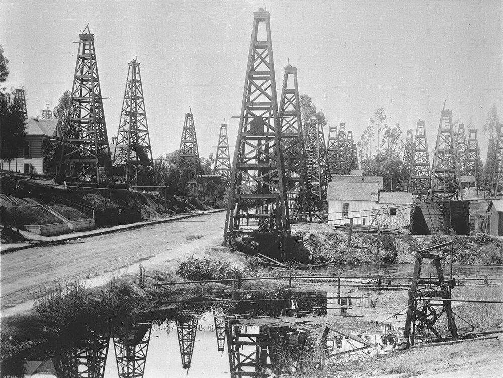 Historiskt svartvitt foto av tidiga 1900-talets oljeborrtorn och träderricks nära en vattenkropp