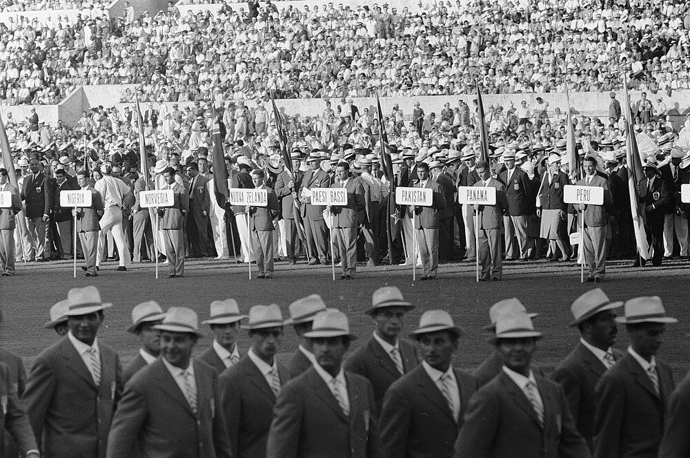 Svartvitt foto av internationella idrottare som marscherar med landskyltar vid ett stort stadionevenemang med publik