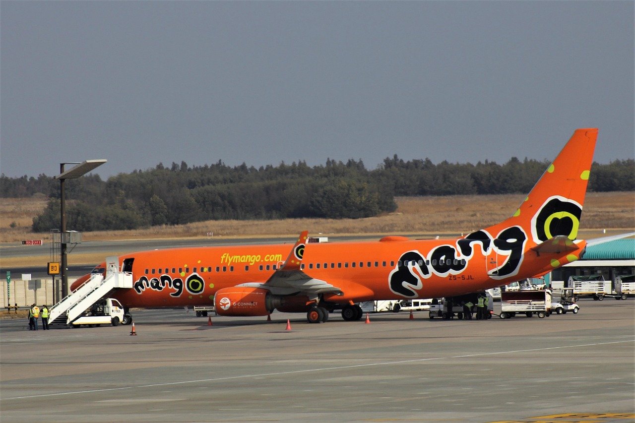 Orange Mango Airlines flygplan parkerat vid flygplatsgate med ombordstigningsstegar och markpersonal som förbereder för avgång
