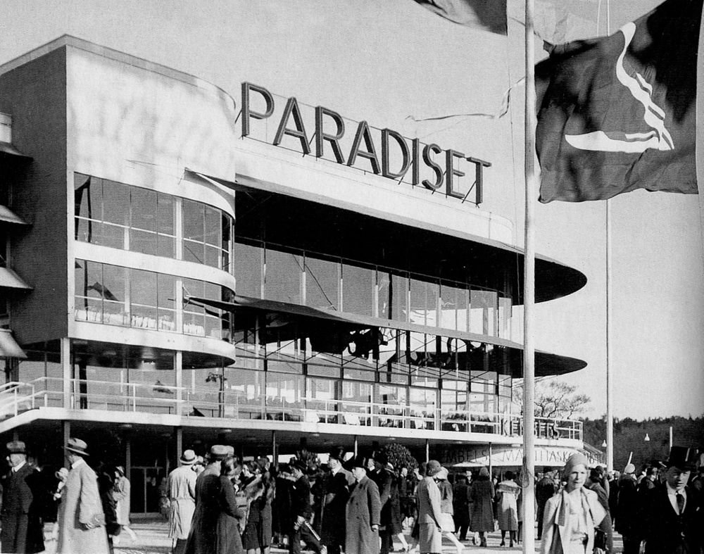 Historiska Paradiset-byggnaden med folkmassa och flaggor Svartvitt foto av en folkmassa utanför Paradiset-byggnaden med stora glasfönster och flaggor i förgrunden