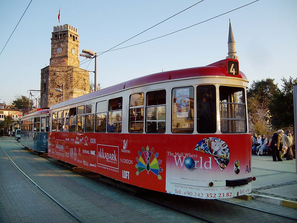 Spårvagn nummer 4 nära klocktornet i Antalya, Turkiet Röd och vit spårvagn nummer 4 vid en station med historiskt klocktorn och turkisk flagga i Antalya, Turkiet
