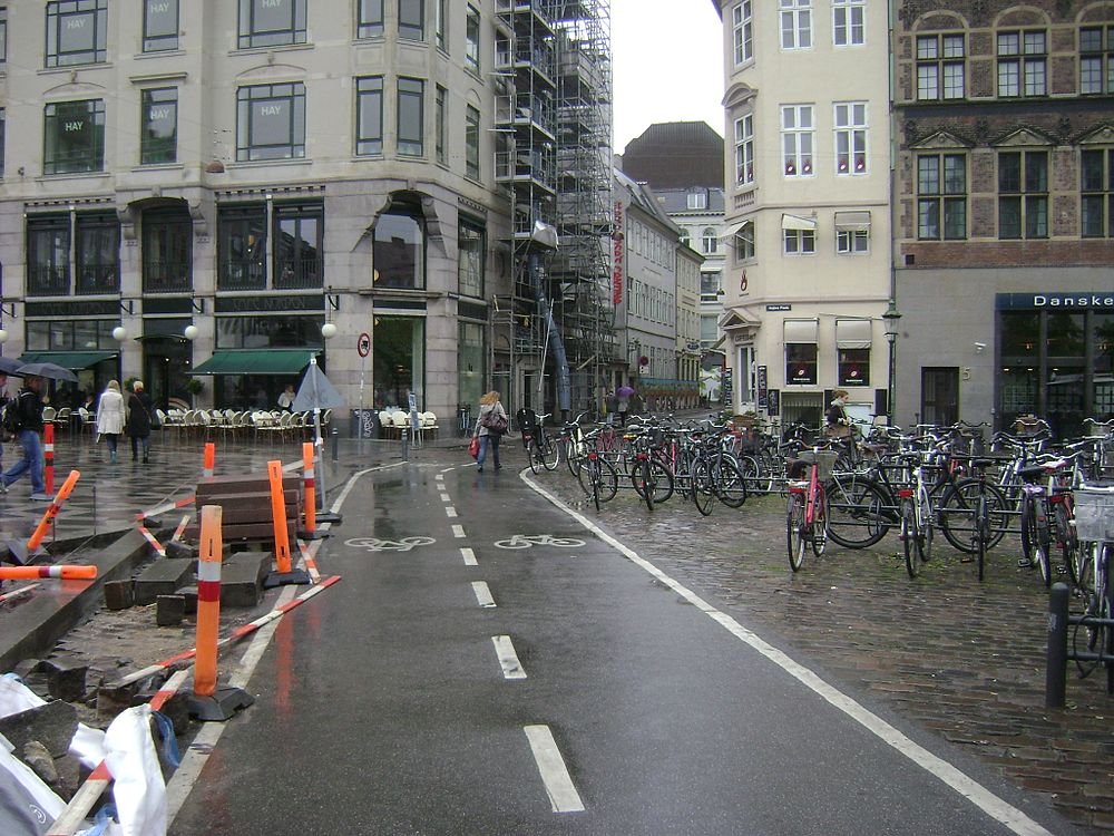 Regnig urban gata med cykelbana och parkerade cyklar Urban gatubild med våt trottoar, cykelbana, många parkerade cyklar och fotgängare nära kaféer och byggnader i regnigt väder