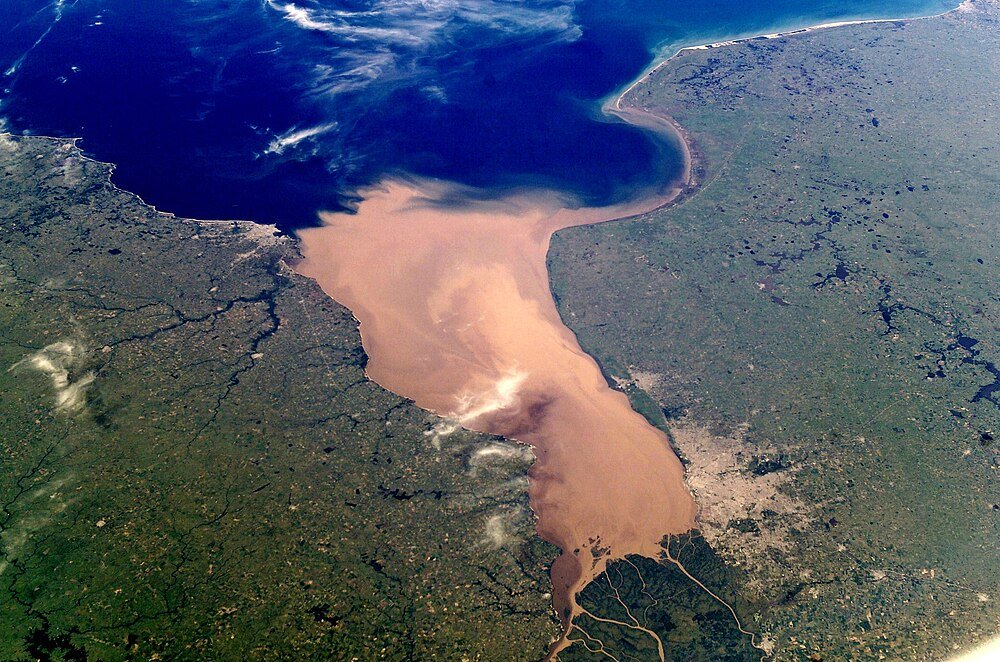 Flygfoto av floddelta som möter havet Flygfoto av ett stort floddelta med lerigt vatten som flyter in i ett djupt blått hav, omgivet av gröna och bruna landområden.