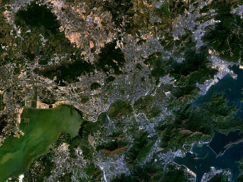 Satellitbild av kuststad och omgivande landskap Satellitvy över en kuststad med urbana områden, gröna berg och vattenområden inklusive en bukt och flod