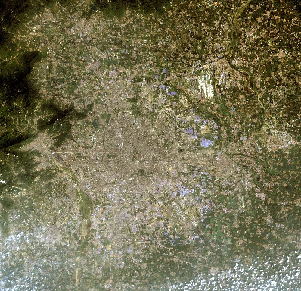 Satellitbild av urban stad och omgivande landskap Satellitvy av en stor urban stad omgiven av jordbruksfält och bergig terräng med spridda moln