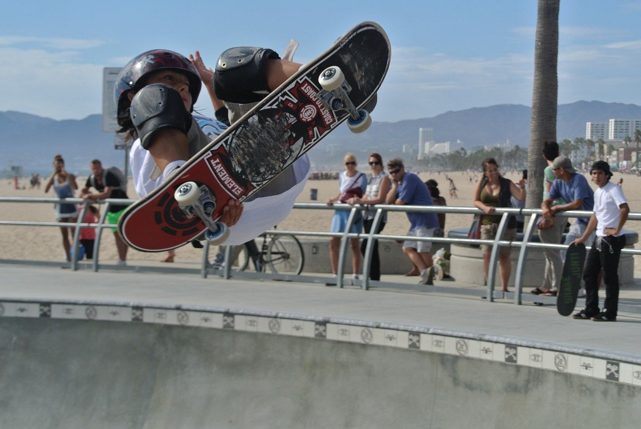 Skateboardåkare som utför ett lufttrick på en solig strand skatepark med åskådare och berg i bakgrunden