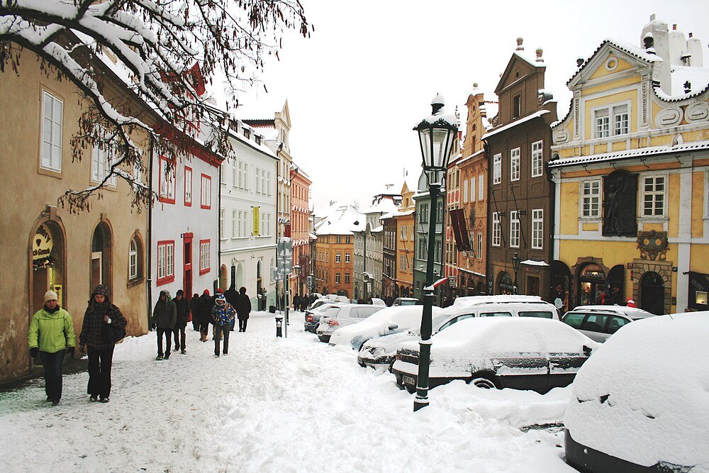 Snötäckt europeisk gata med historiska byggnader Snötäckt gata med parkerade bilar och människor som går nära färgglada historiska byggnader i en europeisk stad under vintern