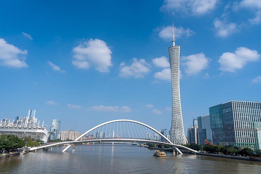 Modern stadsbild med Canton Tower, en vit bågbro över floden och höghus under klarblå himmel