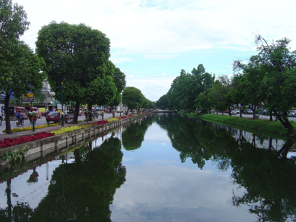 Urban kanal med träd och blommor som speglas i vatten Scenisk urban kanal kantad av frodiga gröna träd och färgglada blommor som speglas i lugnt vatten under en delvis molnig himmel