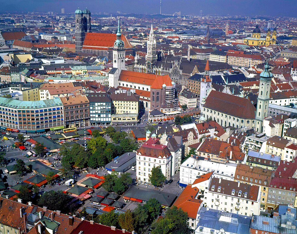 Flygfoto över Münchens stadskärna med historiska byggnader, kyrkor och en livlig utomhusmarknad under klarblå himmel