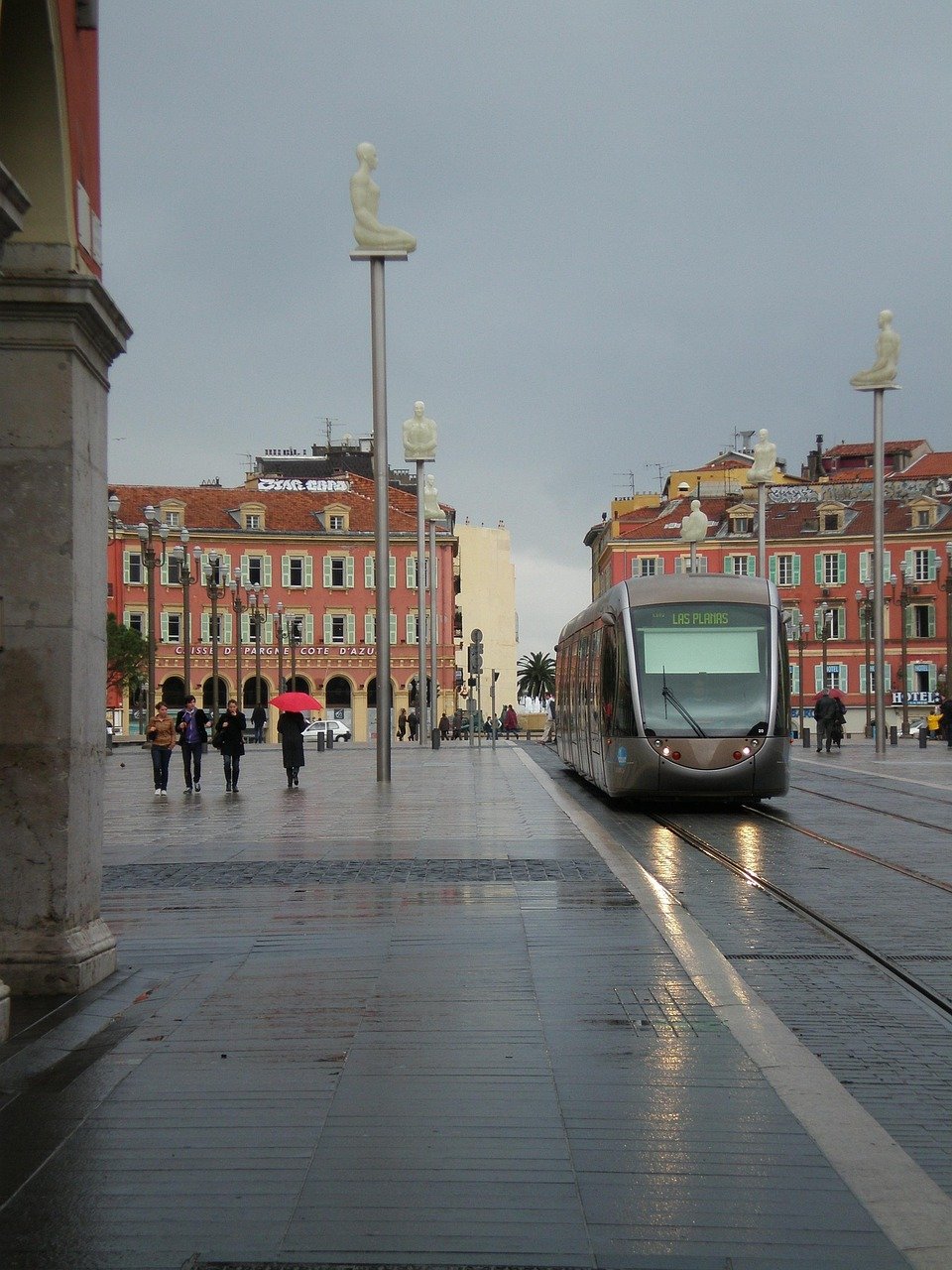 Modern spårvagn på våt gata med unika sittande människoskulpturer på stolpar i ett europeiskt stadstorg