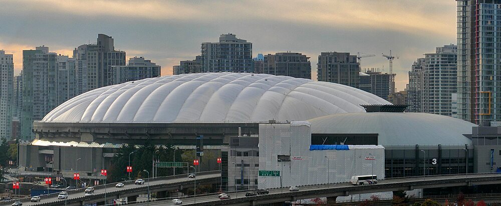Vit kupolformad stadion med stadssilhuett och motorväg Stor vit kupolformad stadion med stadssilhuett och motorvägstrafik i förgrunden under solnedgång