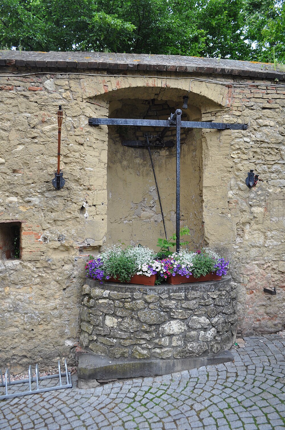 Gammal stenbrunn med färgglada blommor i krukor ovanpå, mot en rustik tegel- och putsad vägg med grönska ovanför