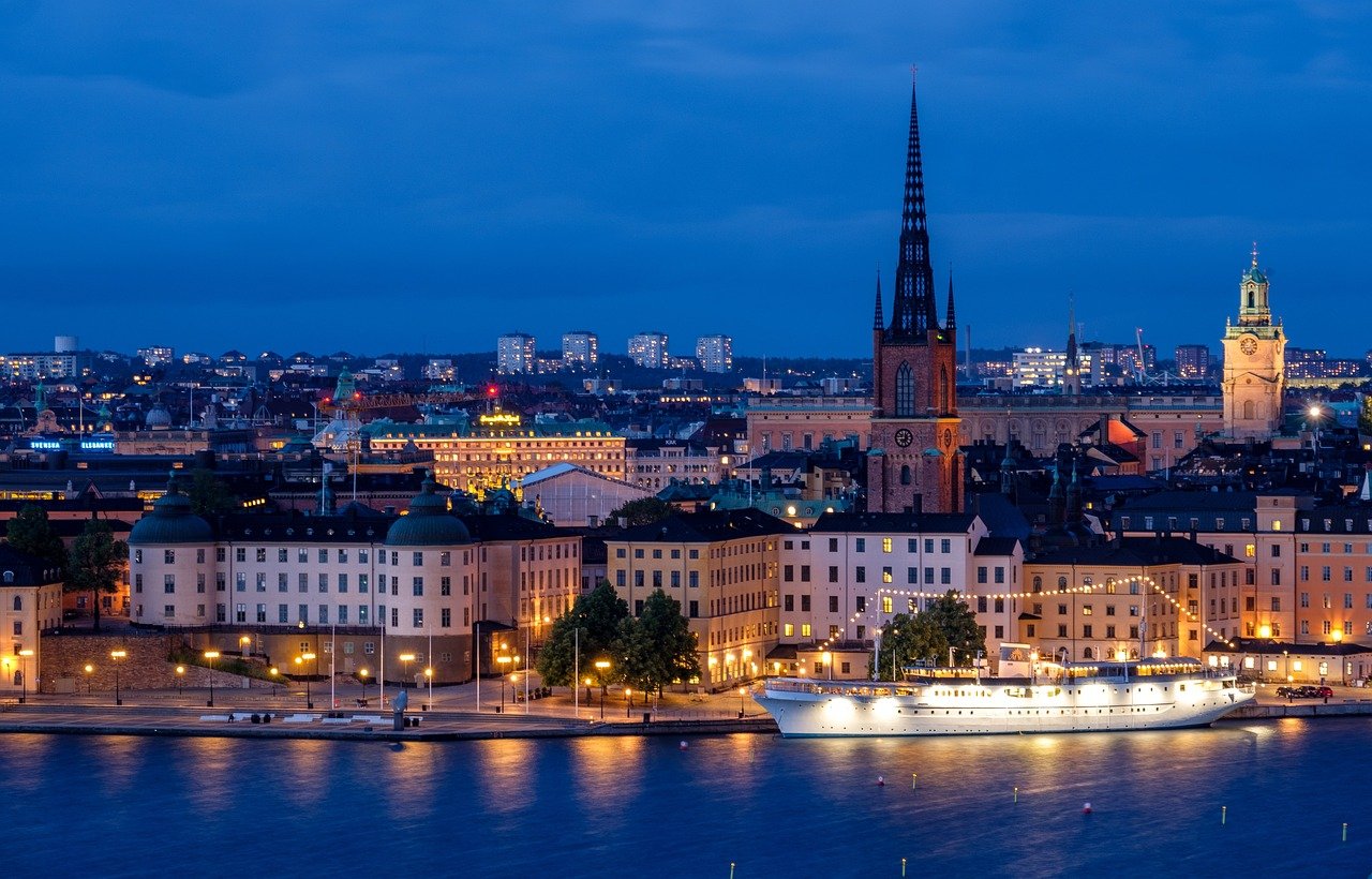 Nattvy över Stockholms vattenfront med upplysta historiska byggnader, kyrkspiror och en förtöjd vit båt som reflekteras i vattnet